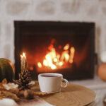 Warm cup of tea, pumpkin, autumn leaves on wooden table on background of burning fireplace. Cozy autumn still life. Thanksgiving and Halloween. Fall hygge